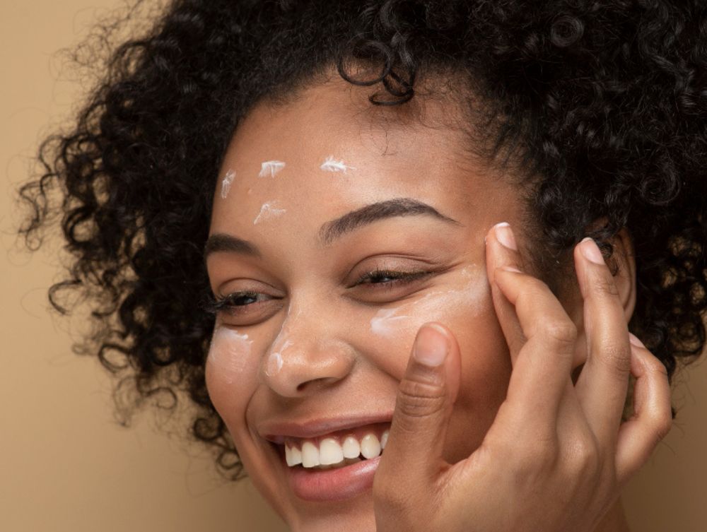 Cuidados diários com a pele: a imagem mostra a modelo sorridente passando um hidratante em seu rosto.